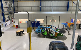 Partially assembled car body on wheeled supports in a clean workshop with exposed components, industrial lighting, and surrounding equipment, used for vehicle testing and development.