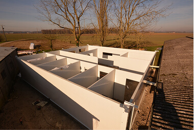 Partially built prefabricated structure with white wall panels forming multiple rooms, located in a rural field during late autumn or winter.