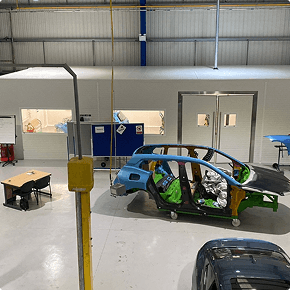 Partially assembled car body shell on wheeled supports in a clean, well‑lit workshop with colored panels, partition wall, and office area in the background.