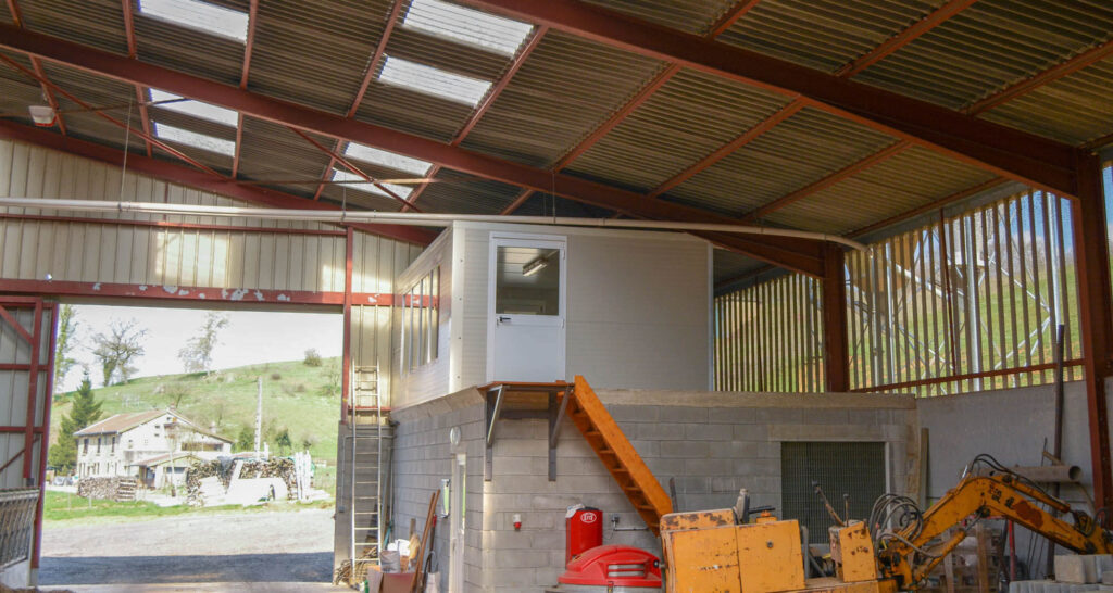 Large metal barn interior with red steel beams, elevated prefabricated office on concrete platform accessed.
