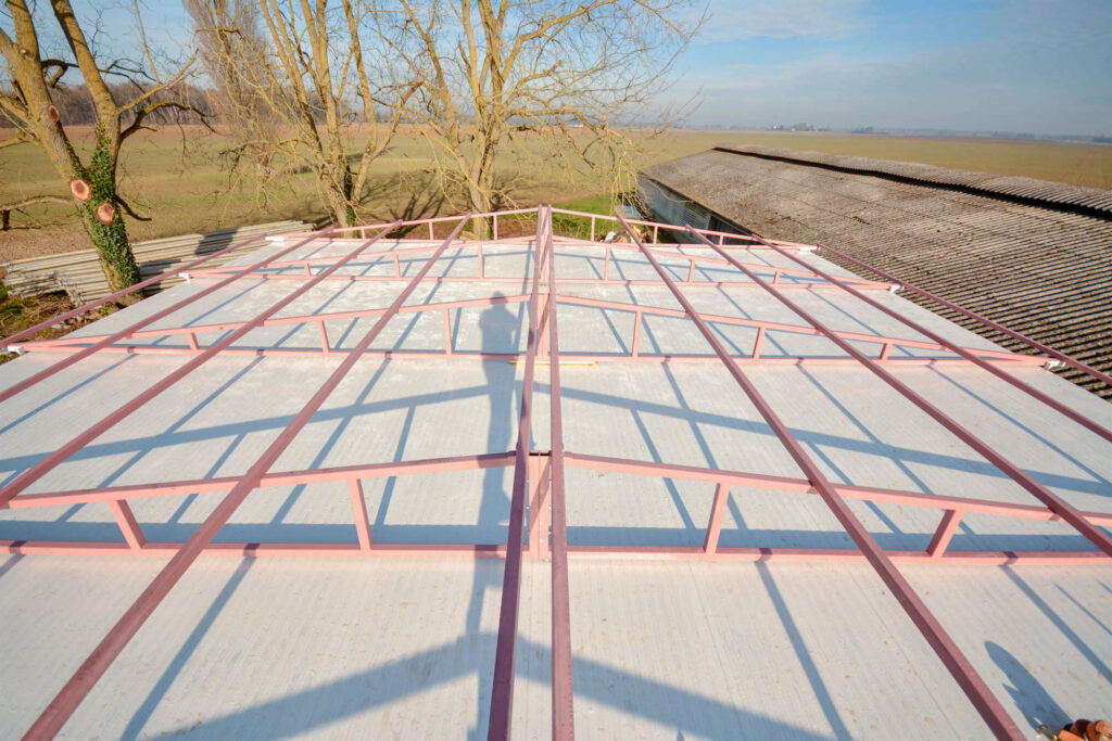 Partially constructed pitched roof with pink metal beams in grid pattern, light surface panels beneath, and rural farmland with trees in the background.
