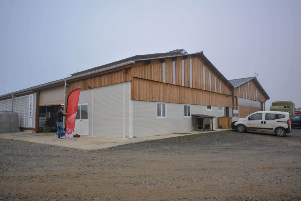 Large industrial farm building with wooden and metal siding, sloped corrugated roof, concrete yard, vehicles parked nearby, and a red Lely banner on a foggy day.