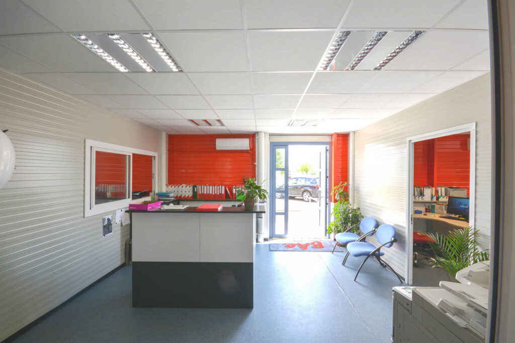 Reception desk with files, folders, and plant, shelves with binders, orange accent panels, visitor chairs, and glass entrance door letting in natural light.
