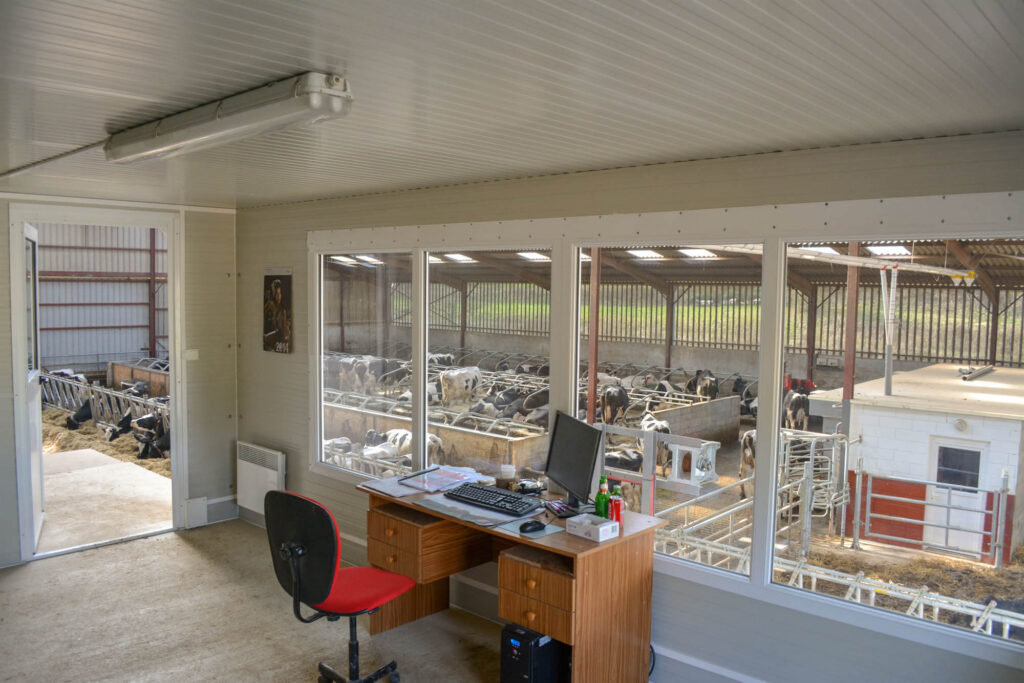 Office with a desk and computer overlooks a large barn filled with cows through wide windows; barn and cattle visible outside.