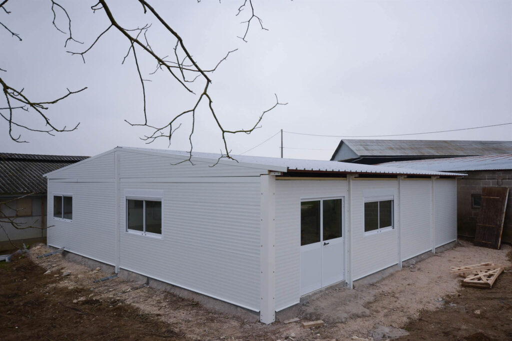 White modular building with corrugated metal walls, sloped roof, multiple windows, and double door, set on concrete foundation with construction materials nearby.