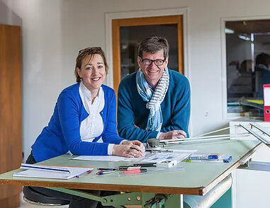 Photograph of two individuals standing behind a large drafting table in a bright office studio, covered with rulers, pens, papers, and mechanical drafting tools, smiling while engaged in collaborative design or planning work.
